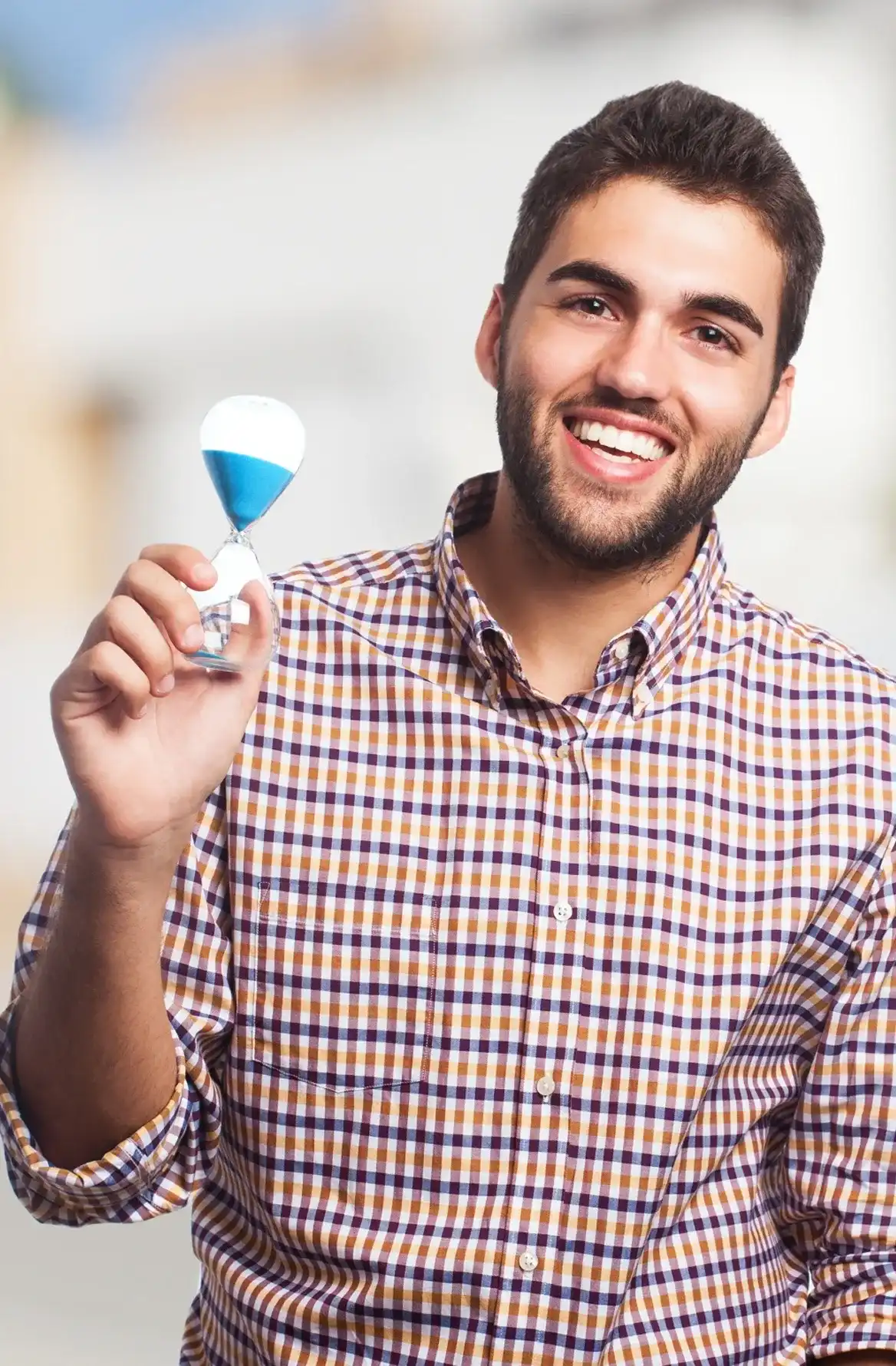 Smiling man in checkered shirt holding a small glass hourglass with blue sand.