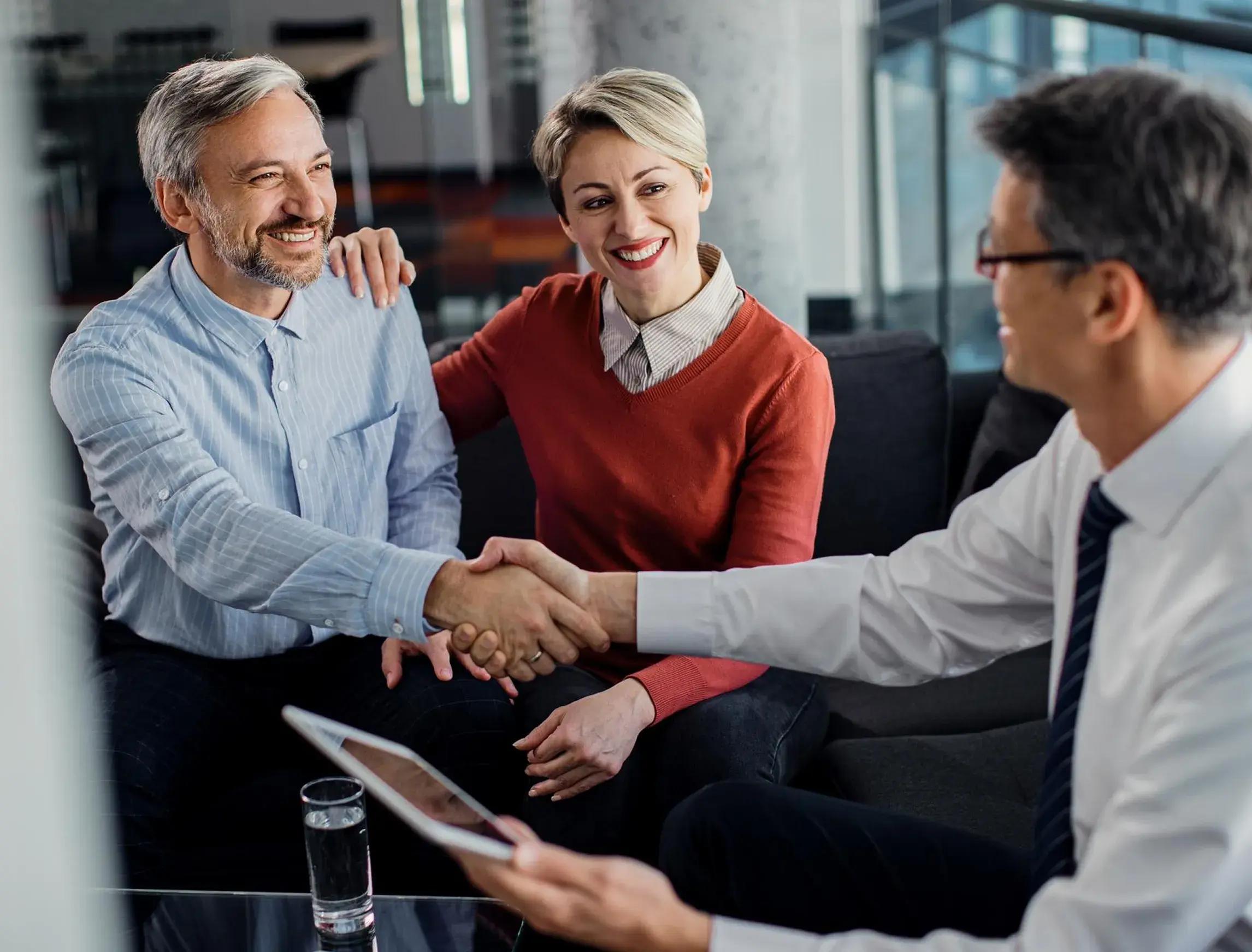 Happy couple shaking hands with a professional man holding a tablet in a modern office setting.
