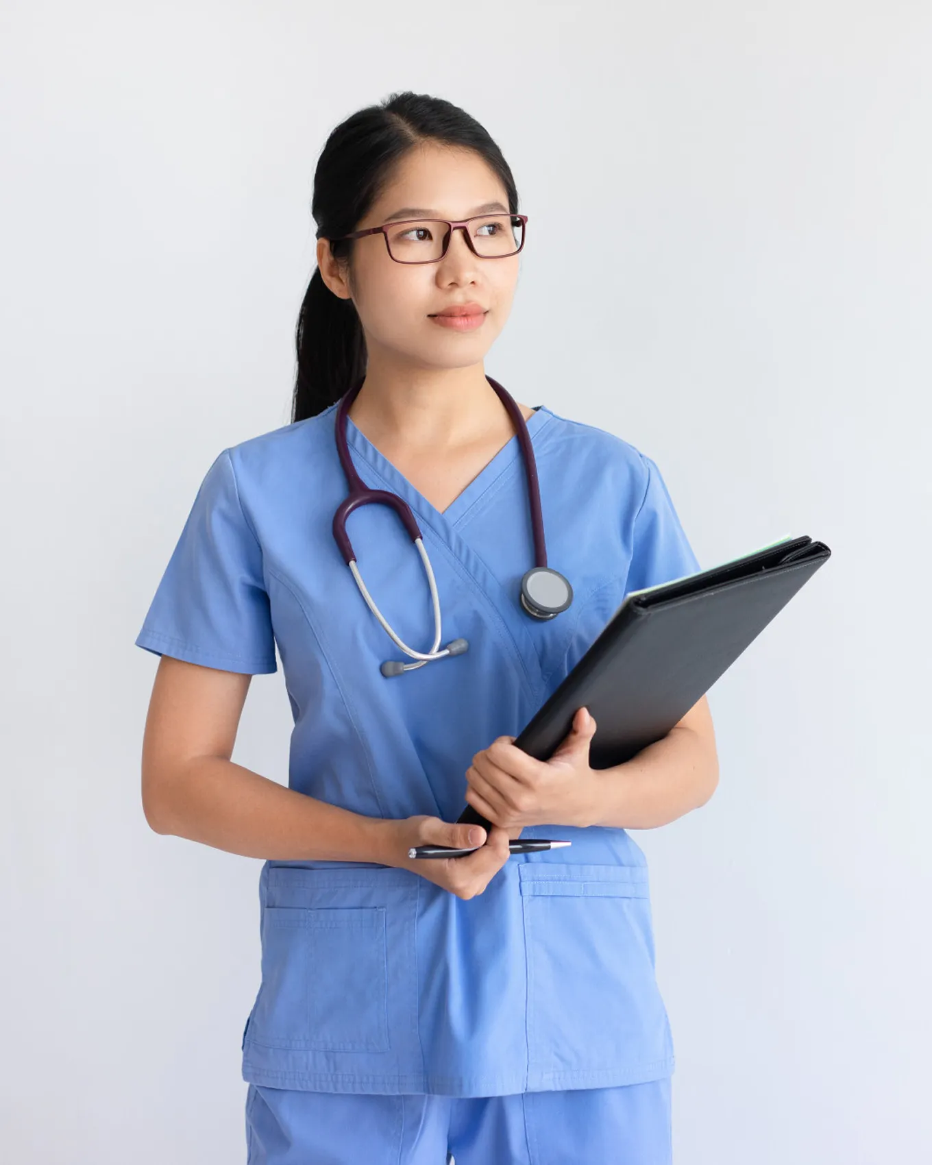 Female healthcare worker in blue scrubs with stethoscope holding black folder and pen.