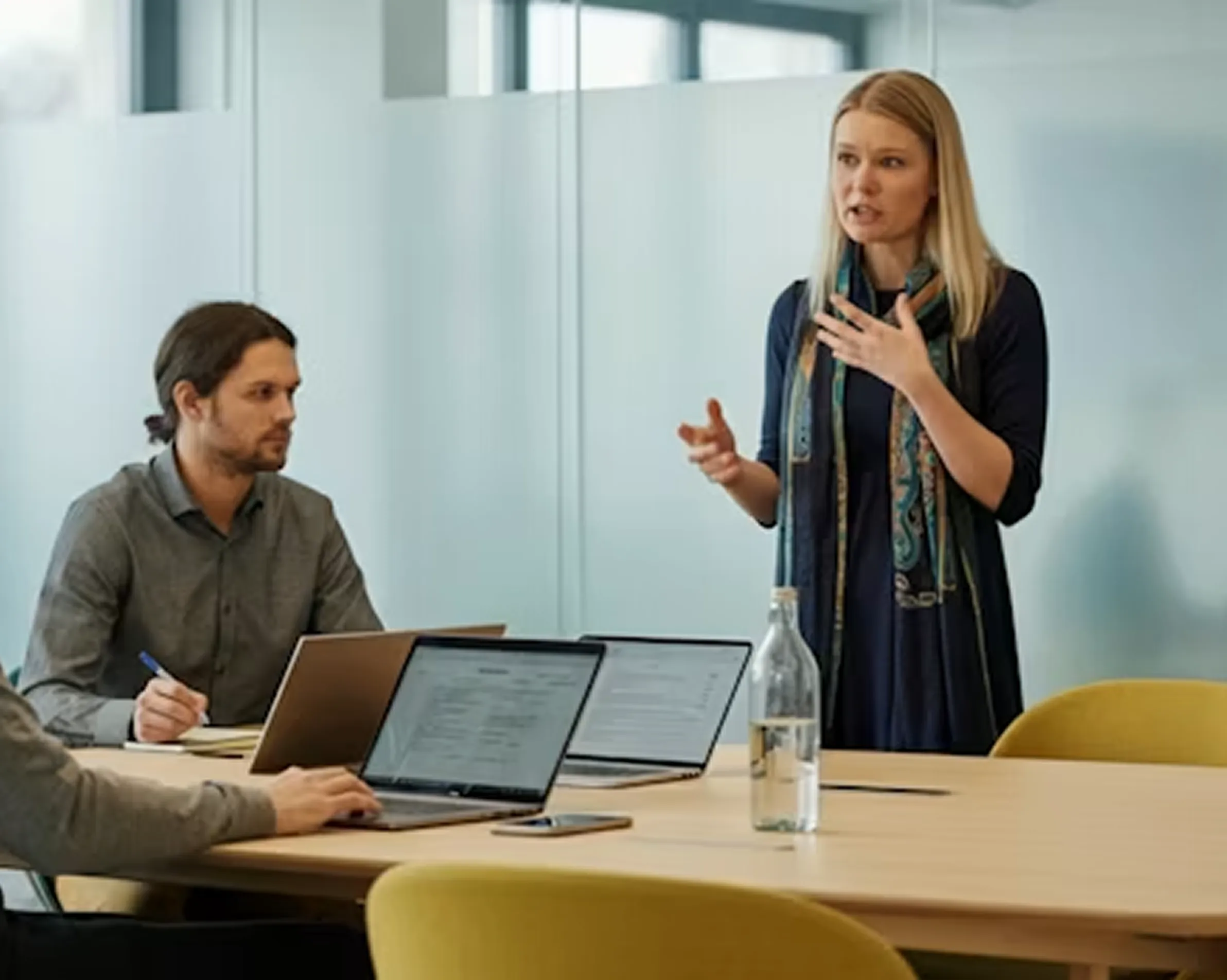 Woman standing and speaking to seated colleagues in office meeting with laptops on table.