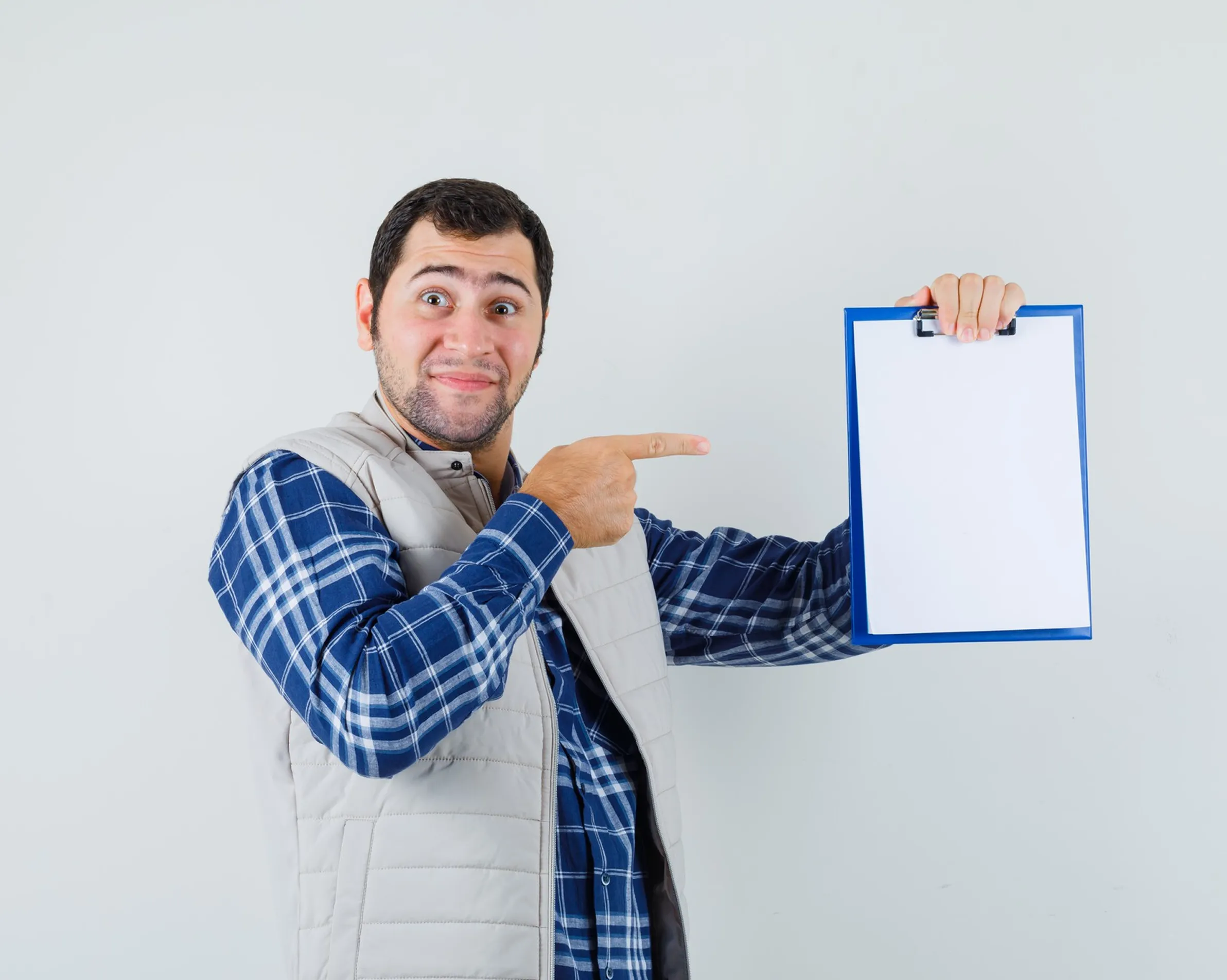 Man in plaid shirt and vest pointing at blank clipboard with a blue border against a white wall.