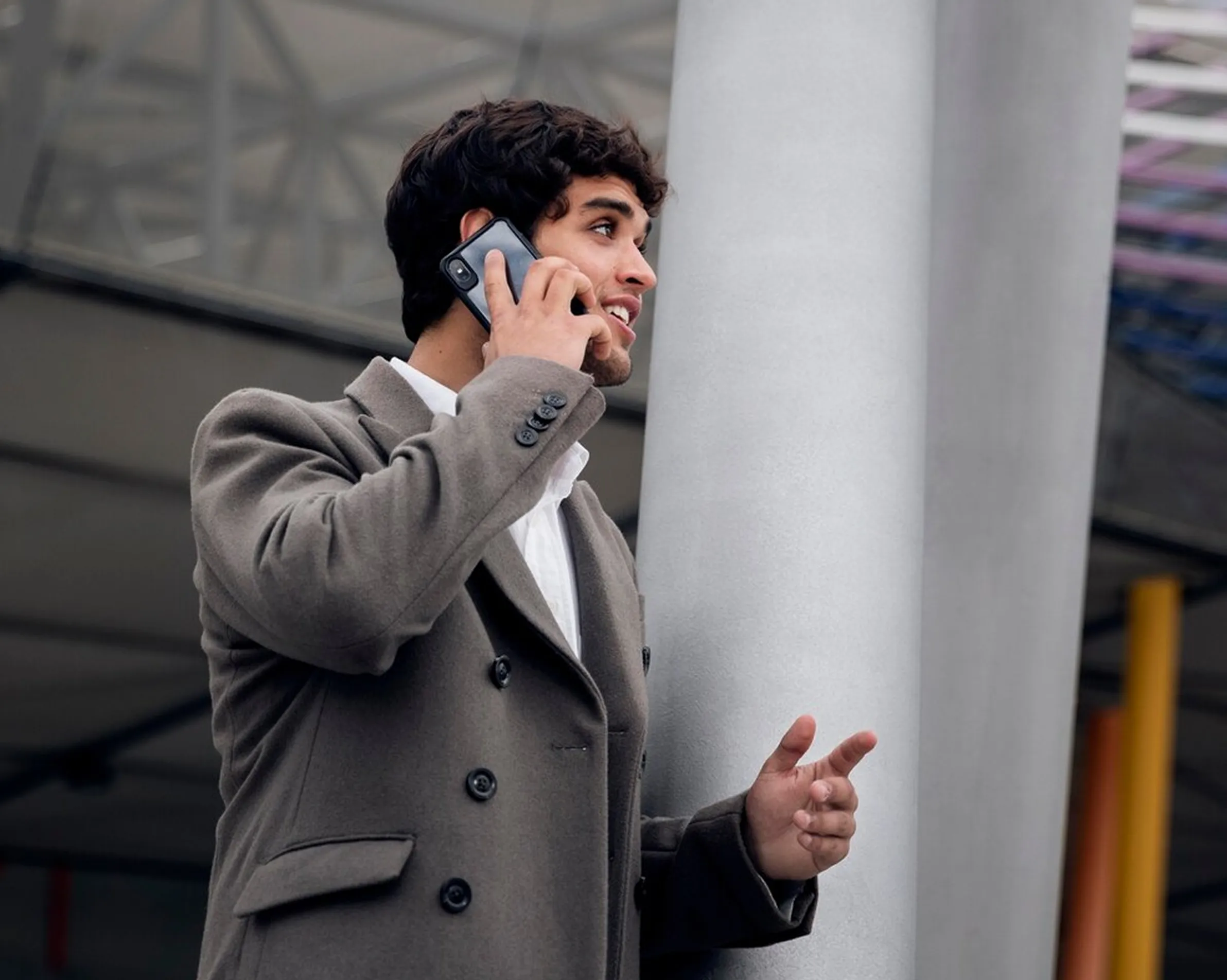 Man in a grey coat talking on a phone, gesturing with his hand near a column in an urban area.