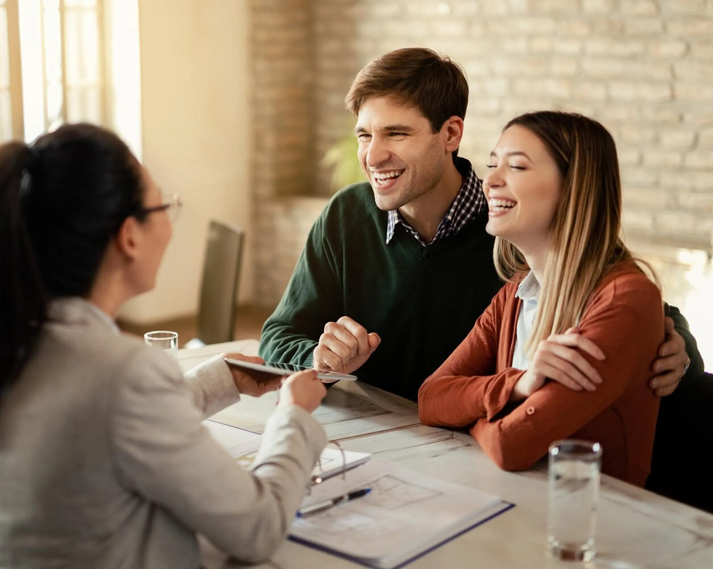 Couple smiling and receiving documents from a woman in glasses at a table with papers and water glasses.