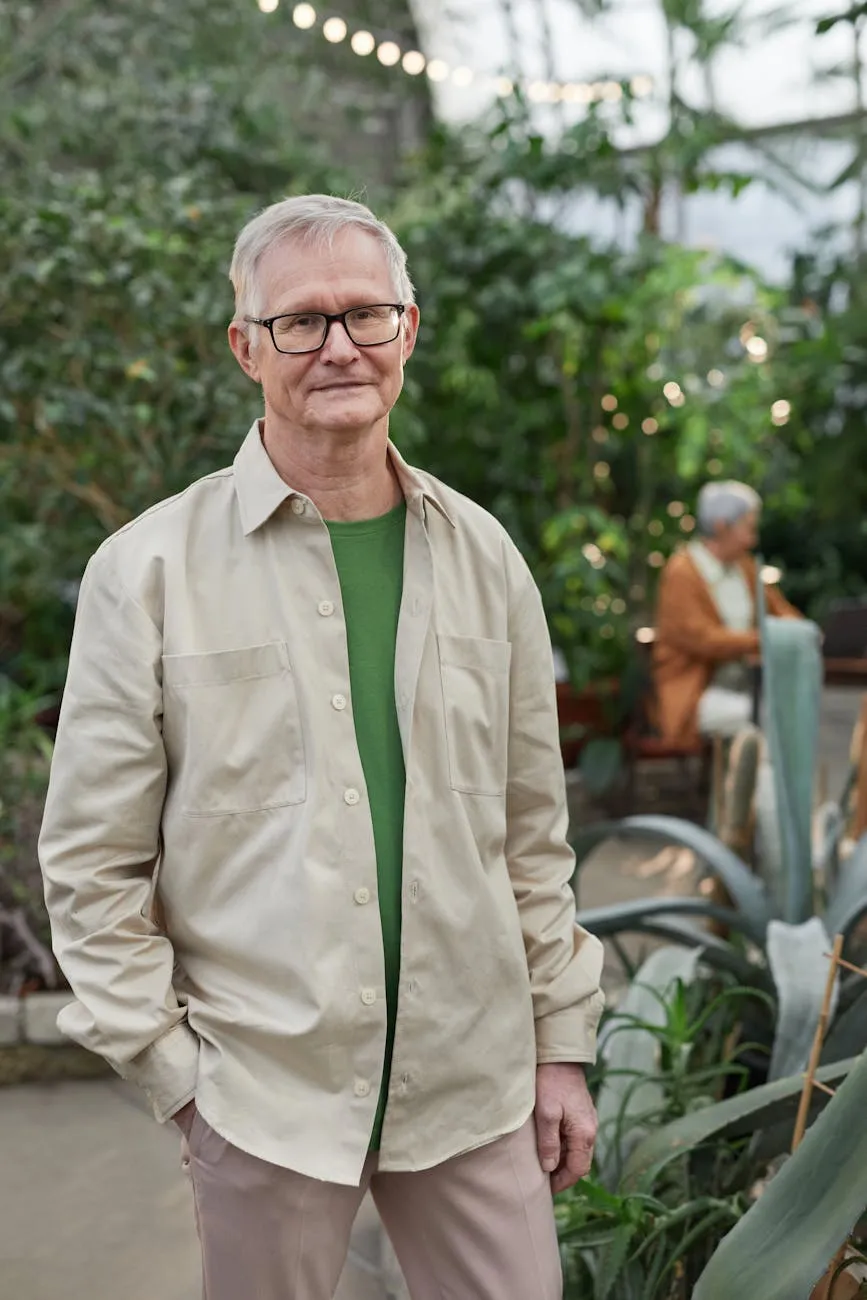 Older man with gray hair and glasses in a beige shirt standing in a greenhouse with plants.