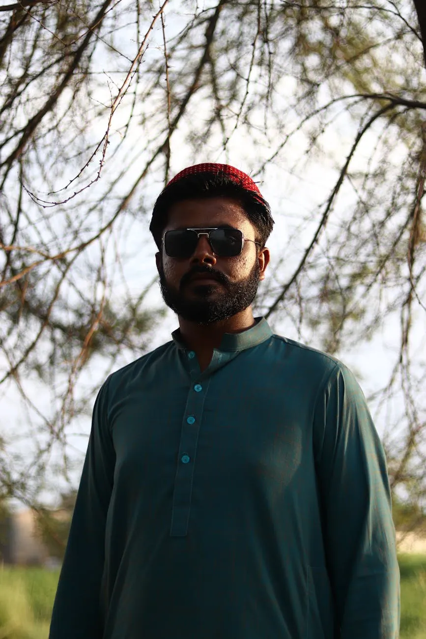 Man with beard wearing sunglasses, a red cap, and a green traditional outfit standing outdoors by bare tree branches.