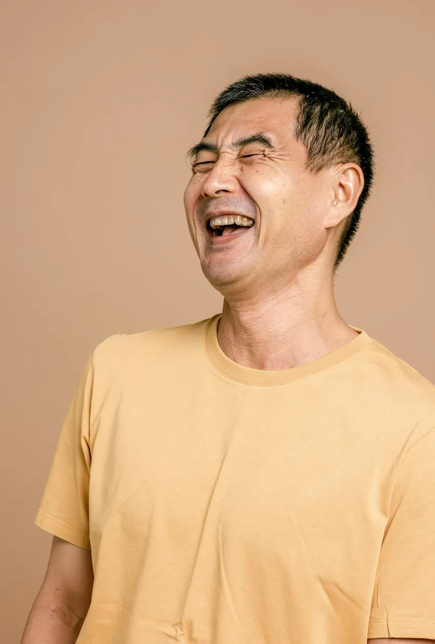Man in a yellow shirt laughing with eyes closed against a beige background.