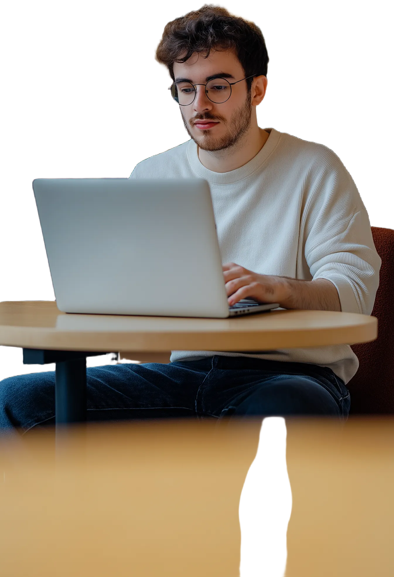 Young man with glasses typing on a laptop at a round wooden table.