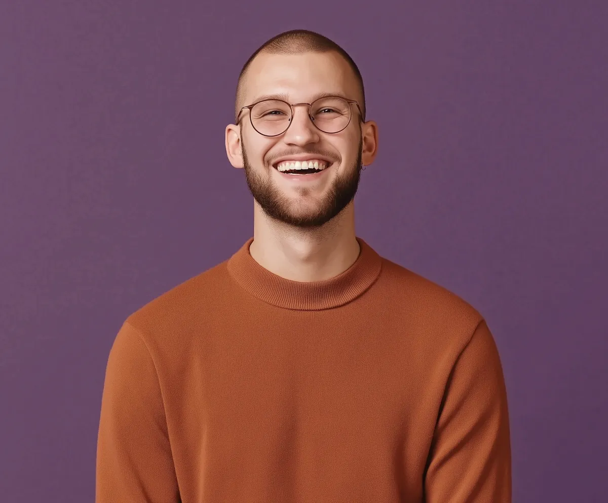 Smiling bearded man wearing round glasses and a brown sweater against a purple background.