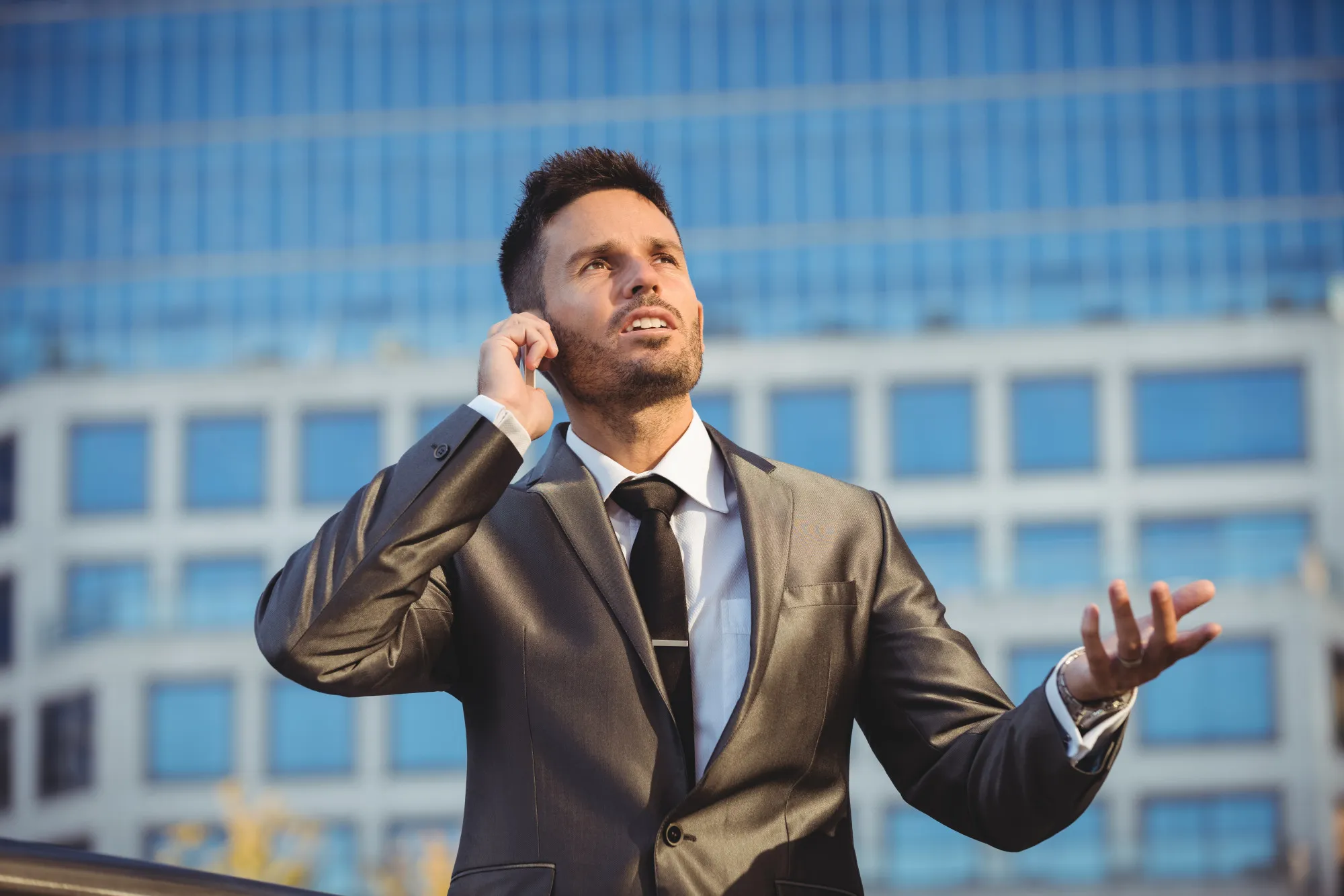 Man in suit talking on phone outdoors in front of a modern glass building, looking upward.