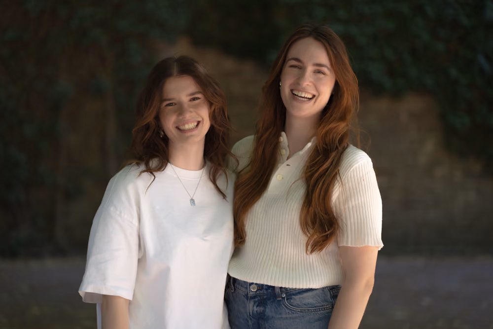 Two smiling young women with long hair standing outdoors in casual white tops, one in a shirt, one in a sweater.
