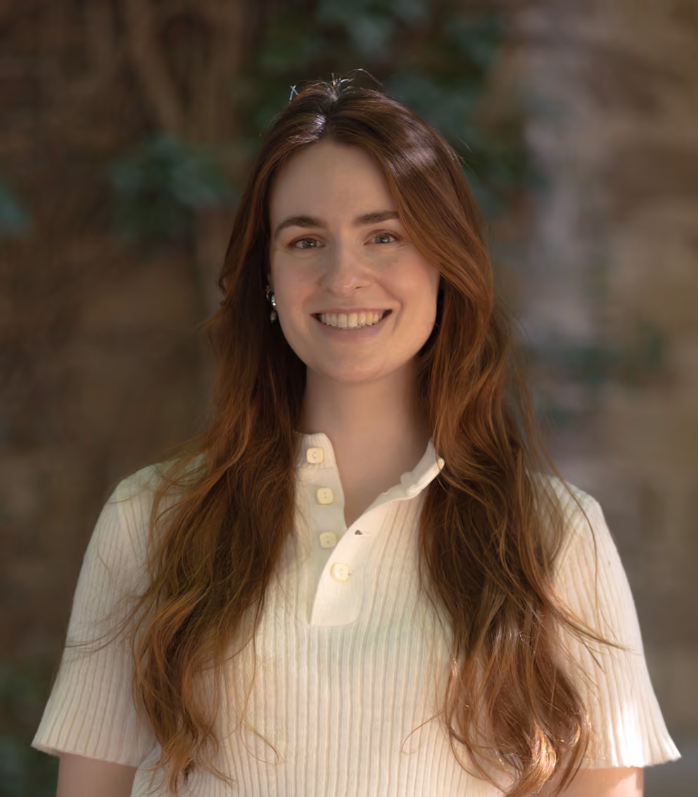 Smiling young woman with long brown hair wearing a white ribbed shirt and blue jeans outdoors.