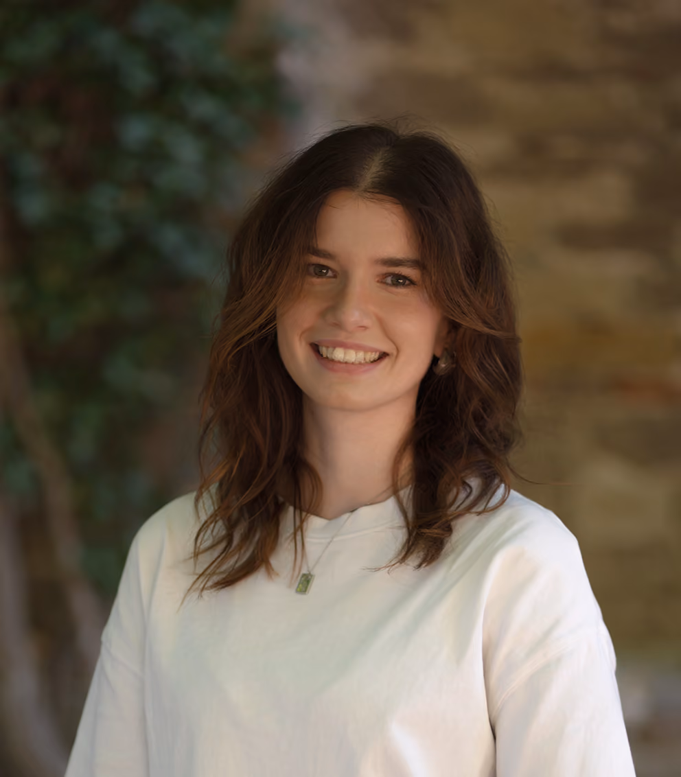 Young woman with wavy brown hair wearing white shirt and jeans smiling outdoors with foliage background.
