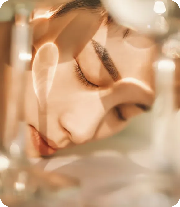 Close-up of a person's face with eyes closed, softly lit with blurred glass objects around.
