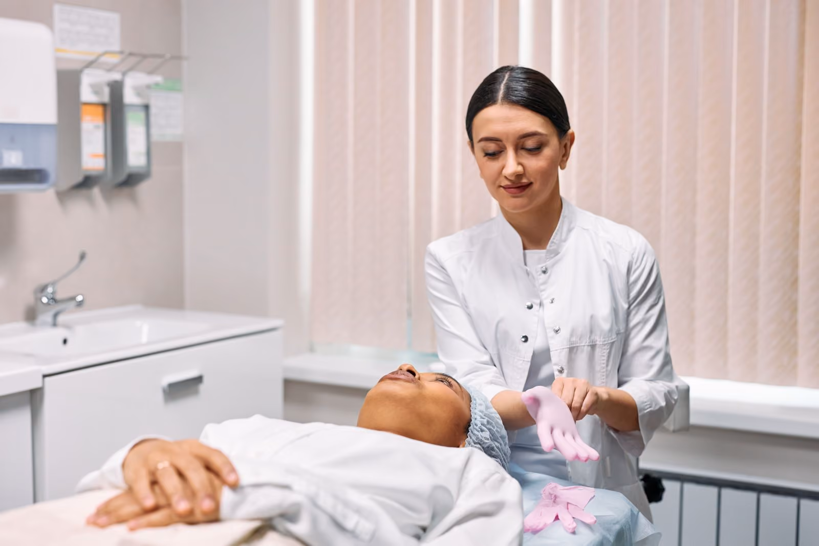 Medical professional putting on pink gloves while patient lies back on exam table in clinic.