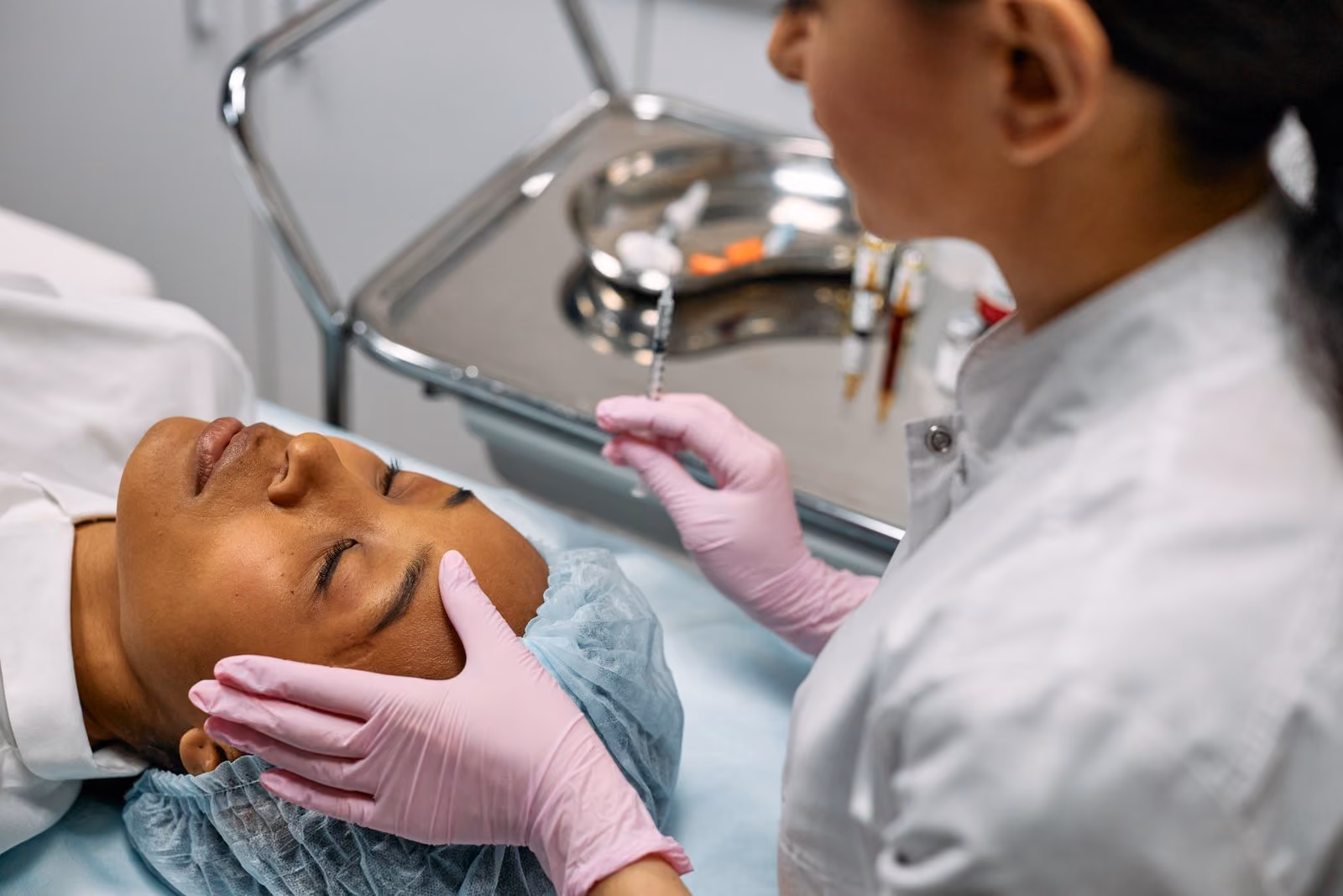 Medical professional in pink gloves prepares syringe near a patient's forehead in a clinical setting.