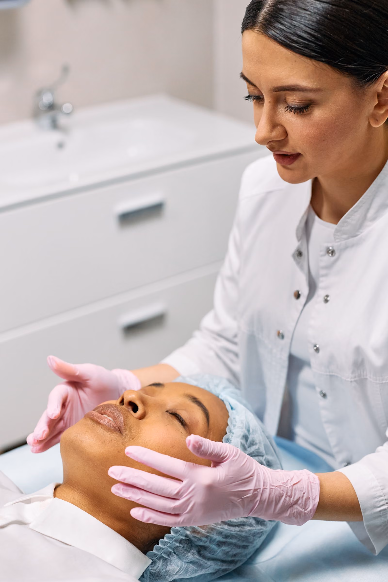 Beautician wearing pink gloves examines face of a woman lying down with eyes closed and hair cover.
