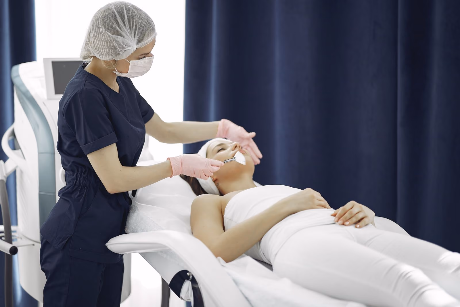 Esthetician in protective gear applies facial treatment to a woman lying on a spa bed.