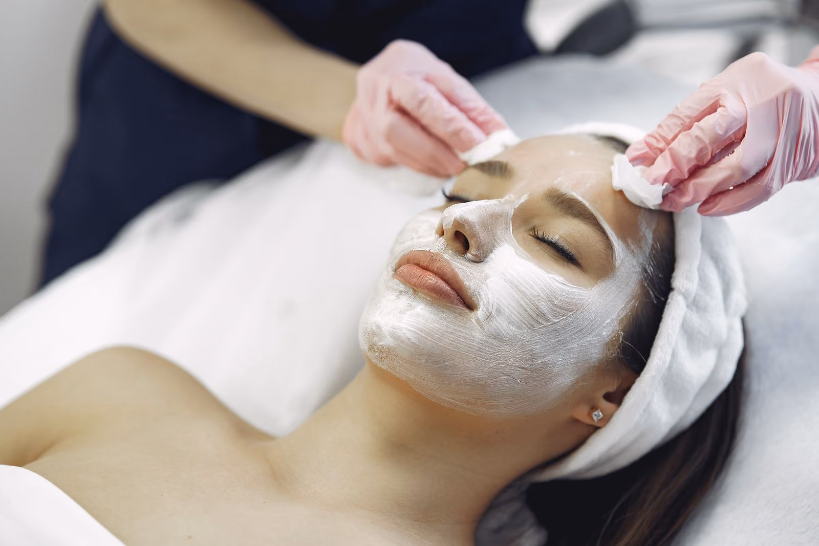 Woman relaxed with white facial mask while a person wearing gloves cleans her forehead with cotton.