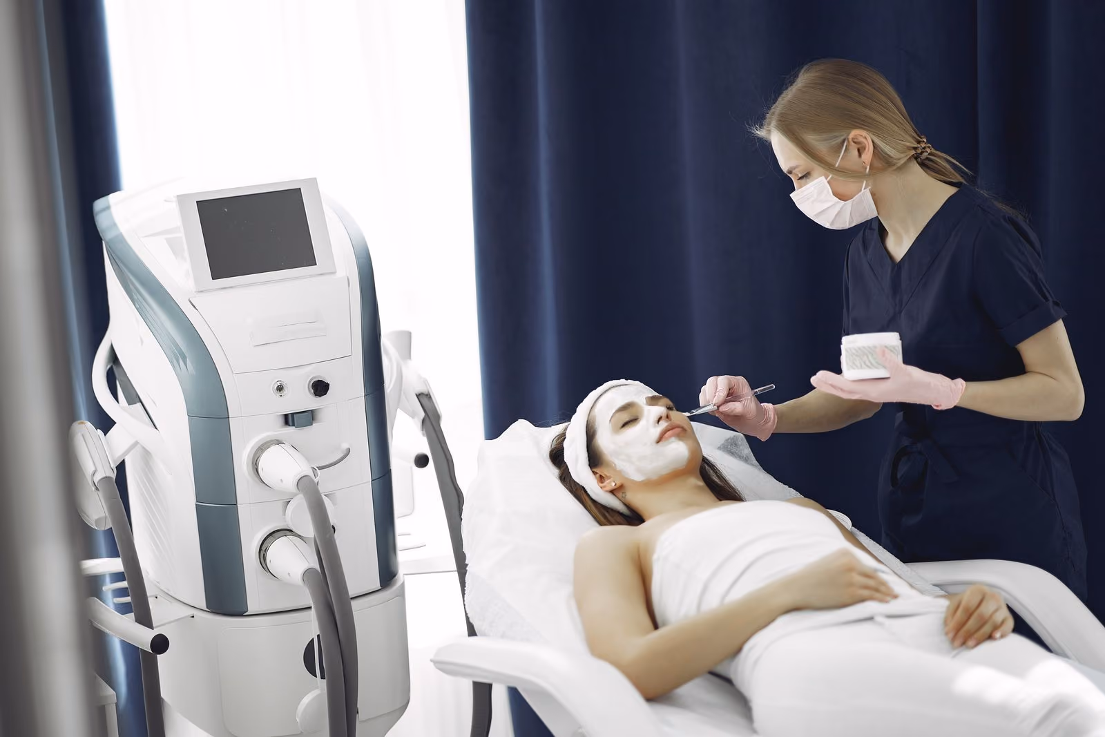 Esthetician wearing gloves and mask applies facial treatment to woman lying on spa bed with face mask.