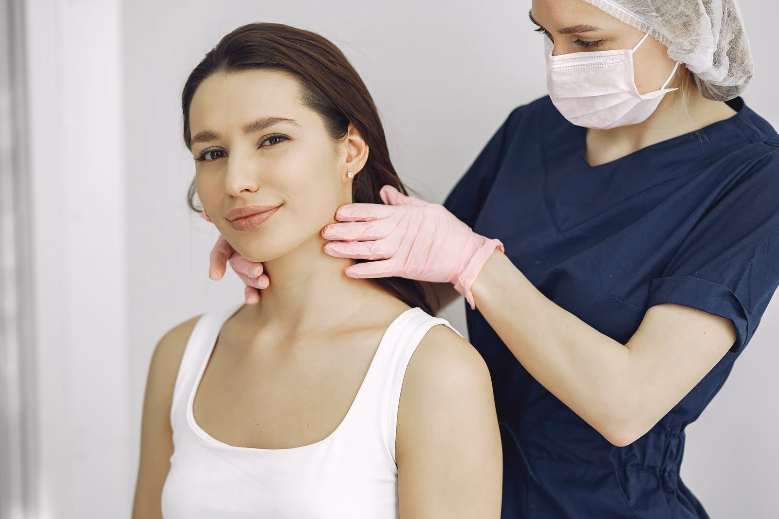Woman in white tank top having her neck examined by a masked healthcare professional wearing pink gloves.