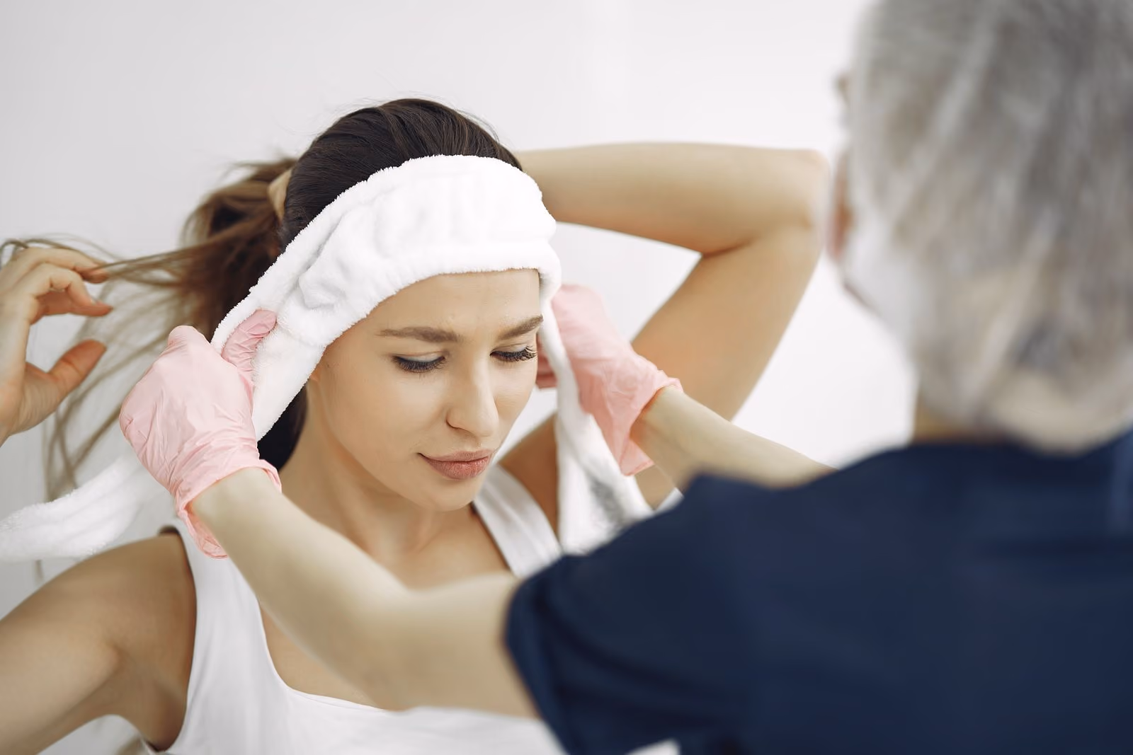 A professional wearing pink gloves puts a white headband on a woman's head in a clinical setting.