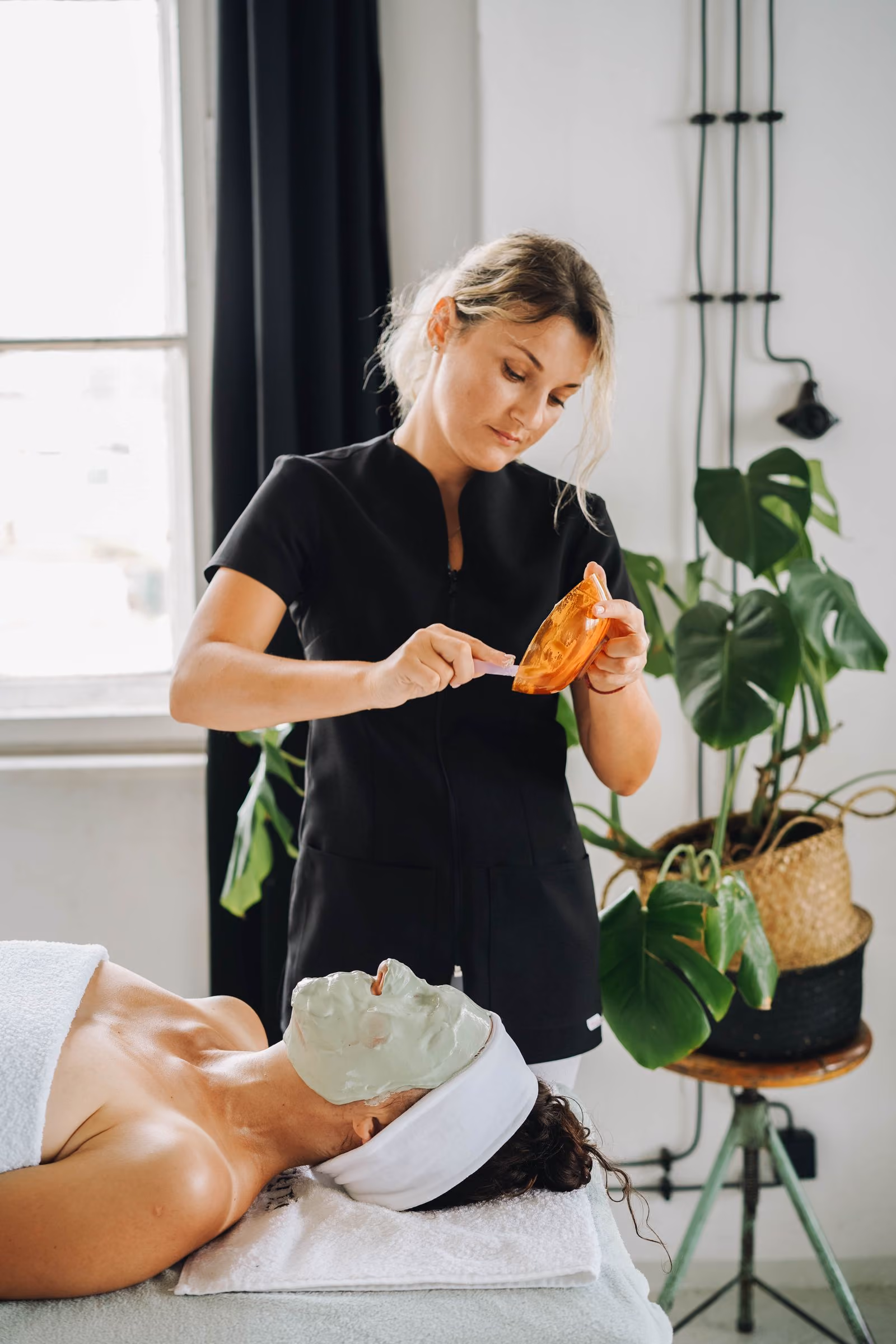 A spa therapist in black applies a green facial mask to a client lying down with a headband.