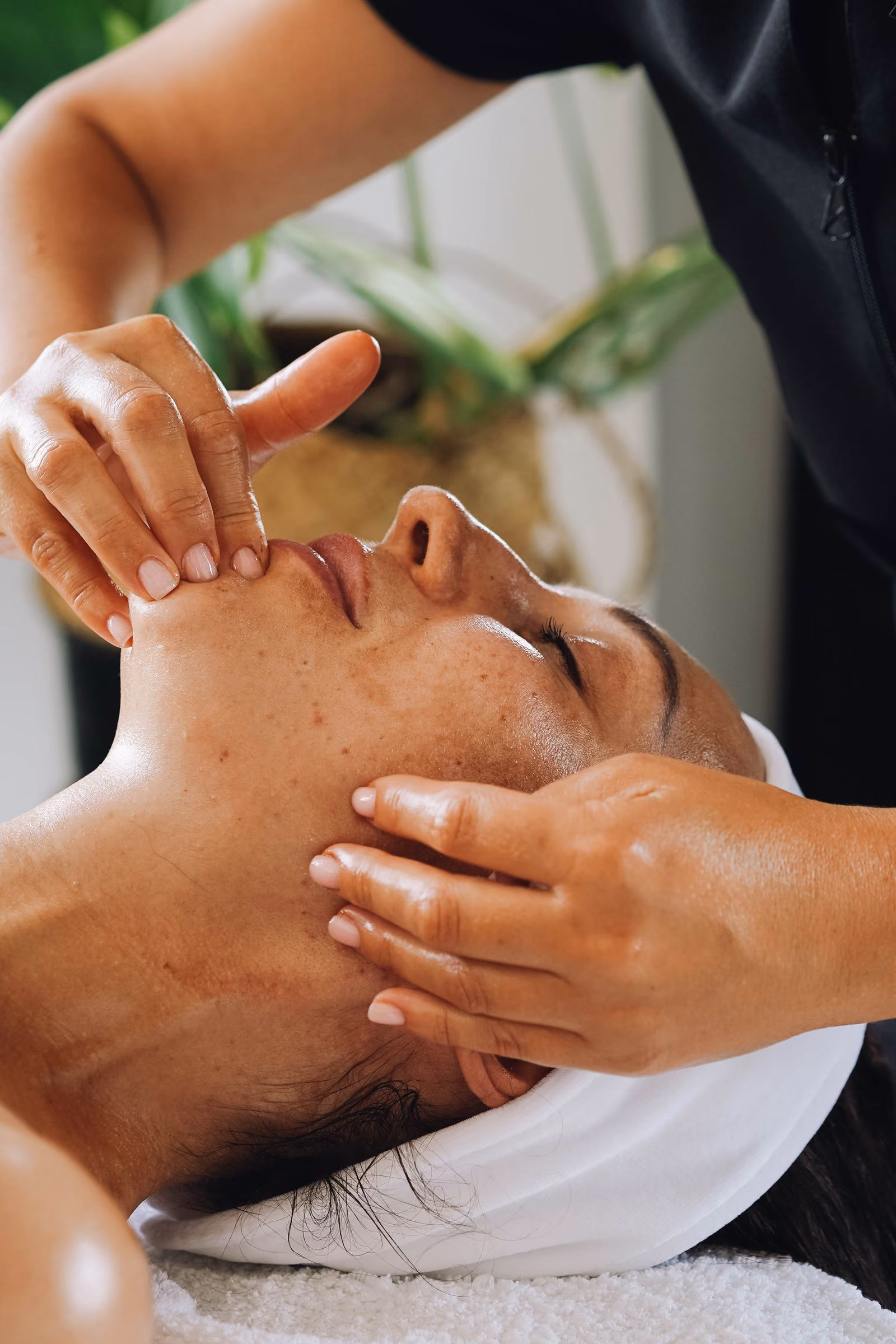 Woman lying down with eyes closed receiving a facial massage from a therapist in a spa setting.