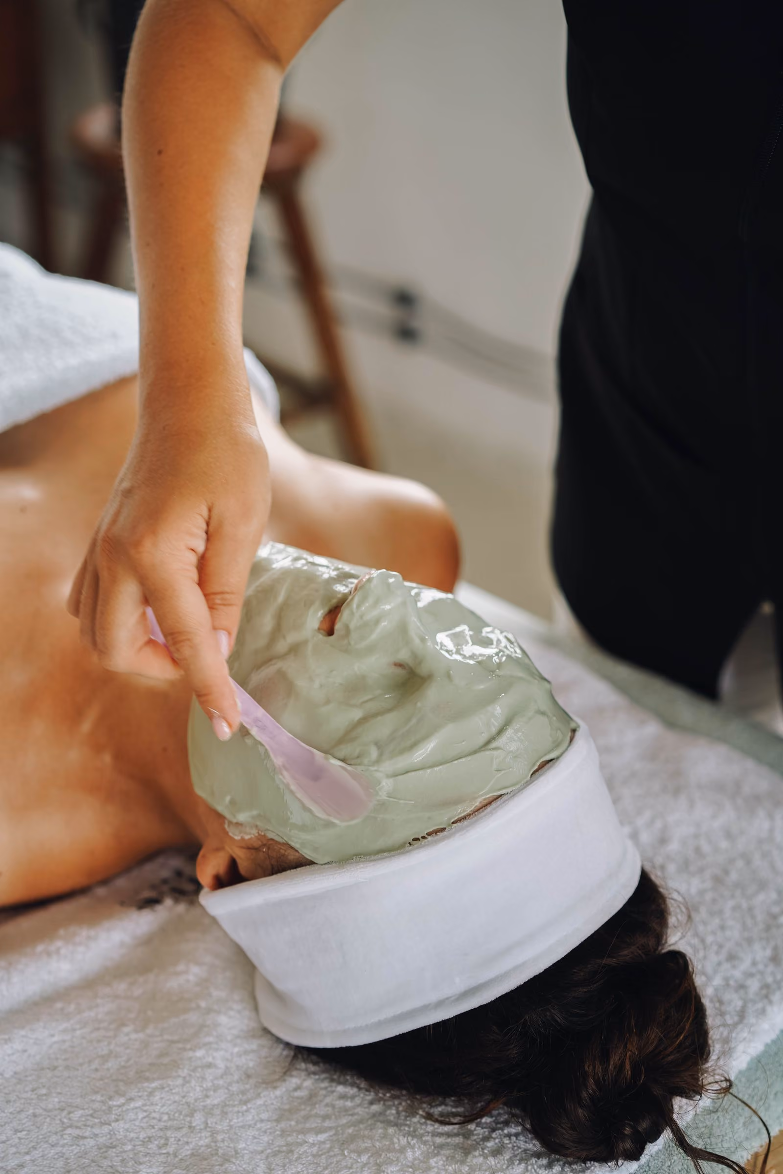 Person lying down while receiving a green facial mask applied with a spatula on their face.