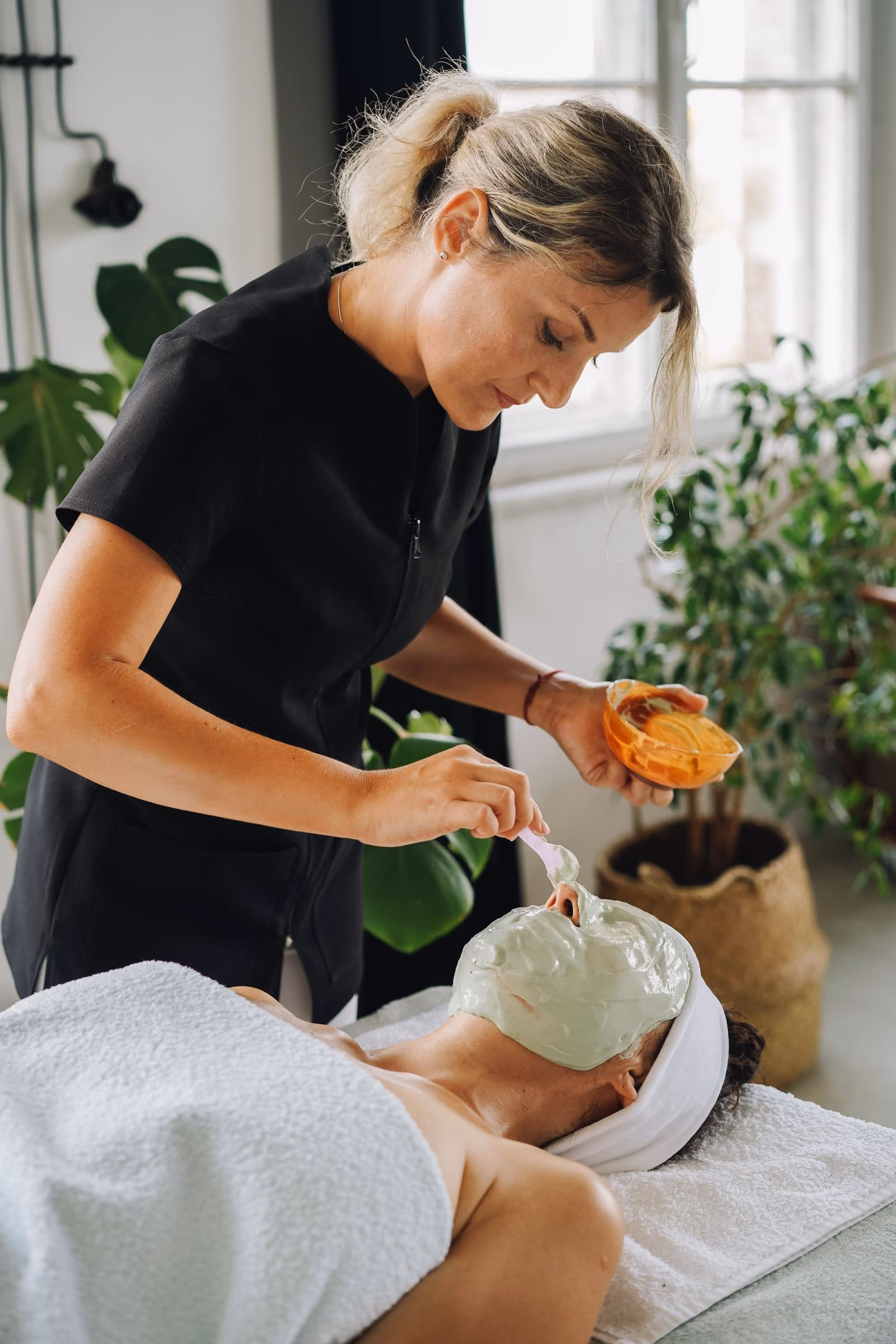 A woman applies a green facial mask on another woman lying down with a white towel and headband.