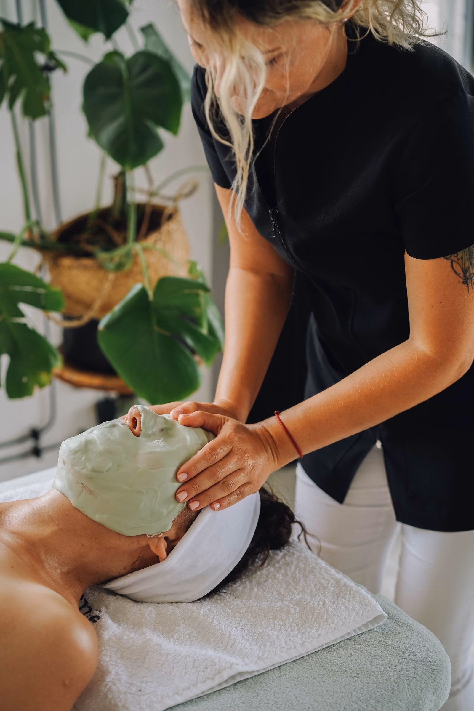 Spa therapist applying a green facial mask to a woman lying on a massage table with a white towel headband.