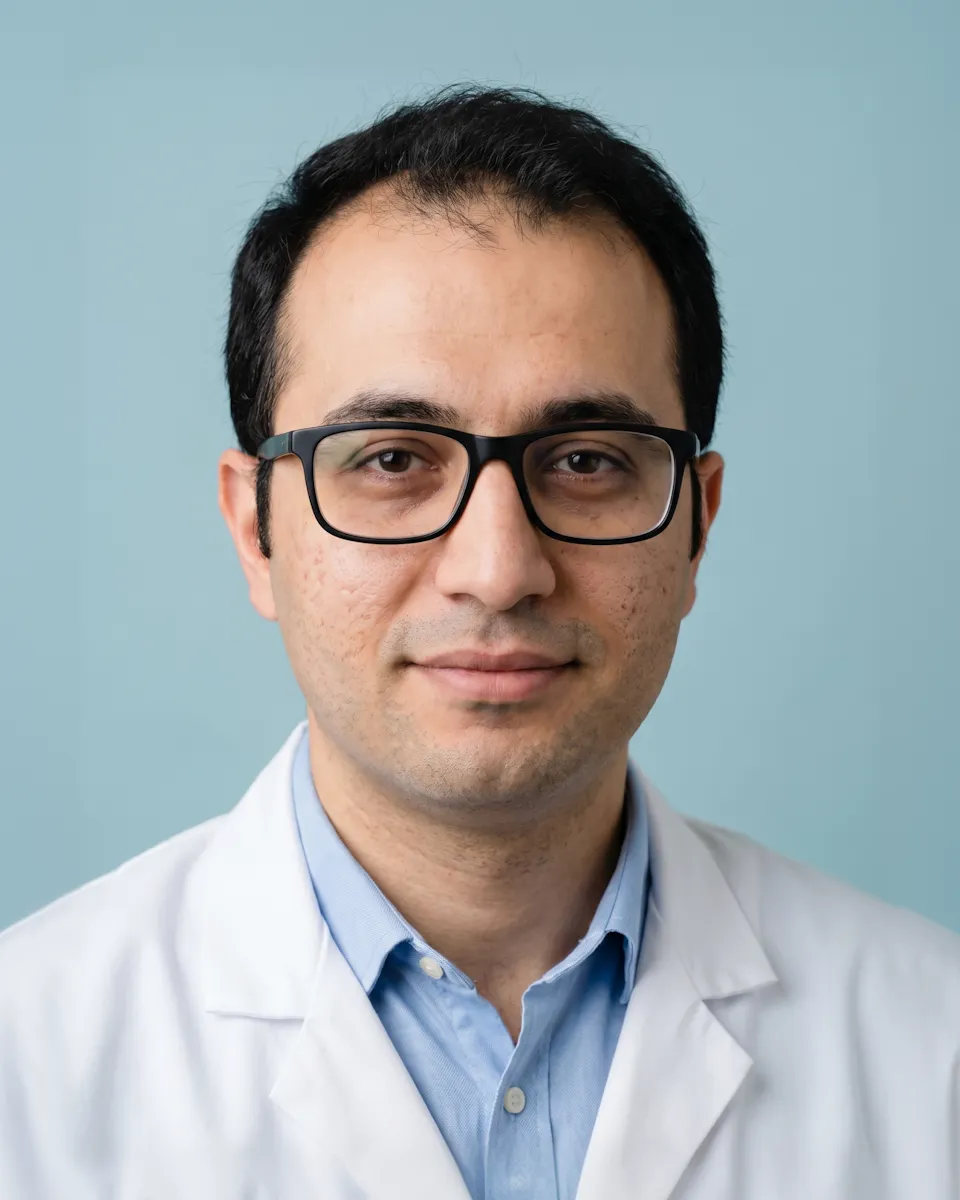 Man with glasses wearing a light blue shirt and white lab coat, with a blue background.