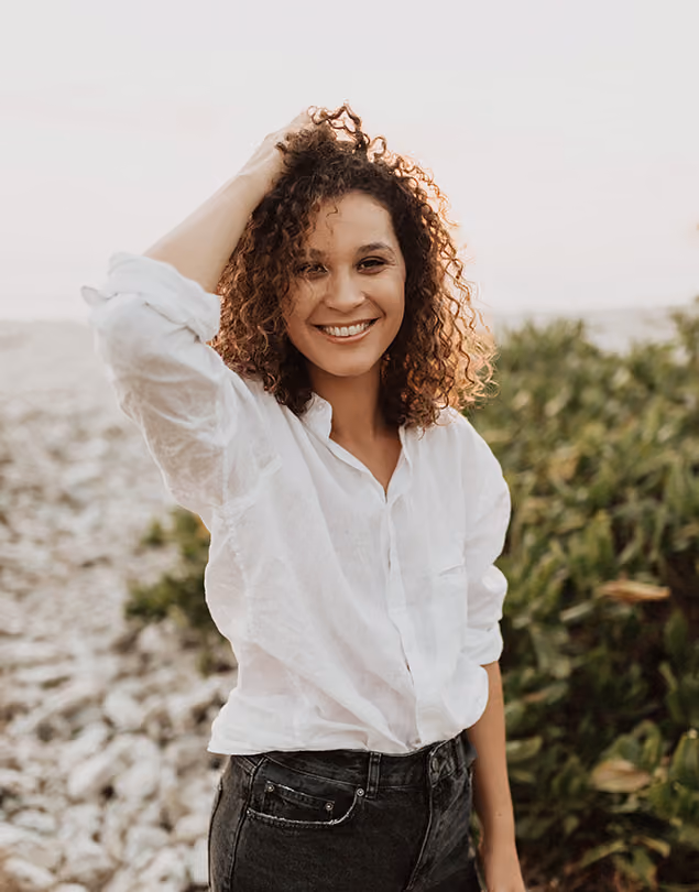 Autoportrait d'Abigail Morellon, photographe à la Réunion. Femme souriante aux cheveux bouclés portant une chemise blanche devant des plantes et des cailloux.