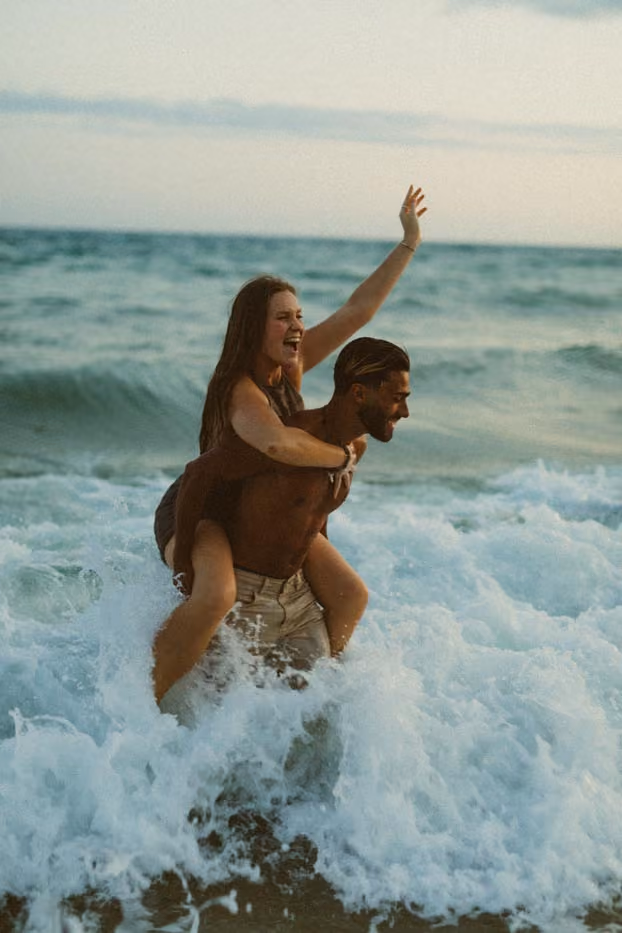 Un homme porte une femme sur ses épaules dans les vagues d'une plage, tous deux souriants.