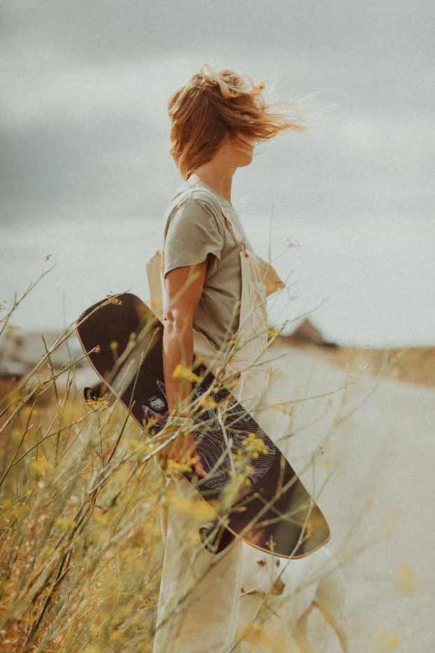 Une femme blonde vue de profil porte son skateboard à la main au milieu des herbes folles, ses cheveux soufflés par le vent.