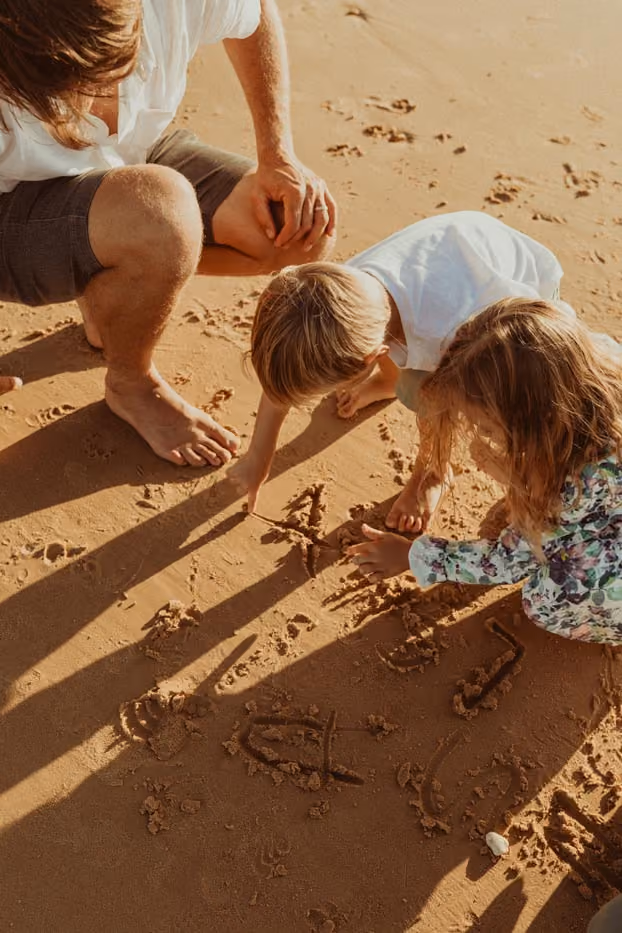Un père et ses deux enfants assis sur le sable chaud dessinent sur la plage avec leurs doigts, capturés en lumière rasante.