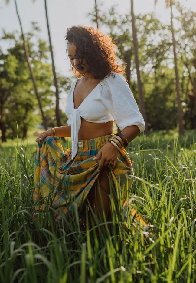 Une femme réunionnaise aux cheveux bouclés se tient au milieu d'un champ de hautes herbes vertes et lumineuses. Elle porte un une tenue créole typique : un crop top blanc noué à l'avant et une jupe à carreaux, avec de grands palmiers en arrière-plan sous un ciel ensoleillé.