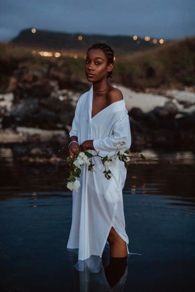 Portrait de Youmna en tunique blanche fluide, debout dans l'eau à la tombée de la nuit avec une branche de fleurs blanches à la main.