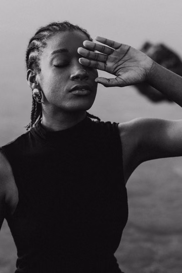 Portrait en noir et blanc d'une femme cachant à moitié ses yeux fermés avec sa main, sur fond de paysage marin.