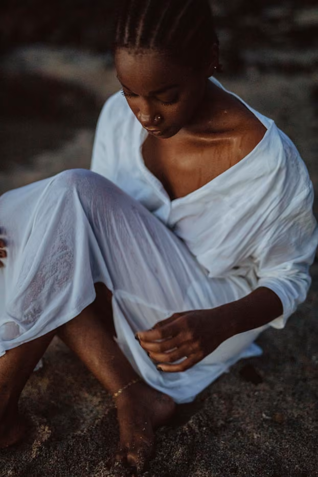 Une femme assise pieds nus sur le sable mouillé en fin de journée, vêtue d'une robe blanche légère et mouillée.