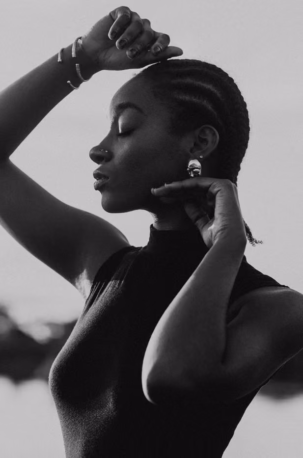 Portrait artistique en noir et blanc de profil d'une femme noire aux cheveux tressés, posant les mains près du visage.
