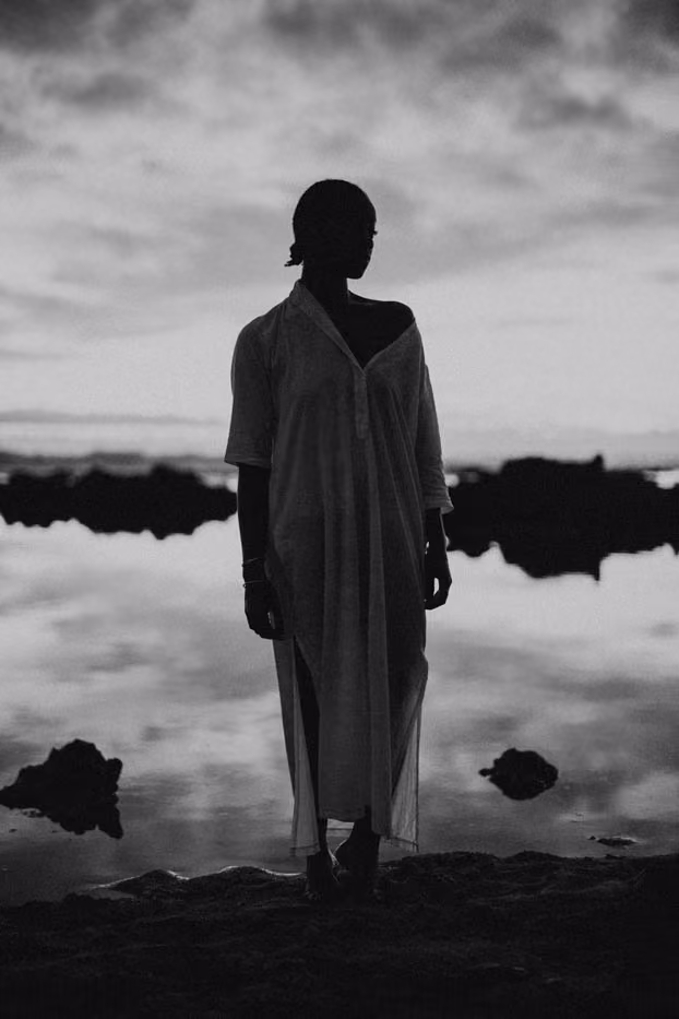 Silhouette en noir et blanc d'une femme debout face à la mer au coucher du soleil, se reflétant sur l'eau calme à Saint-Gilles.
