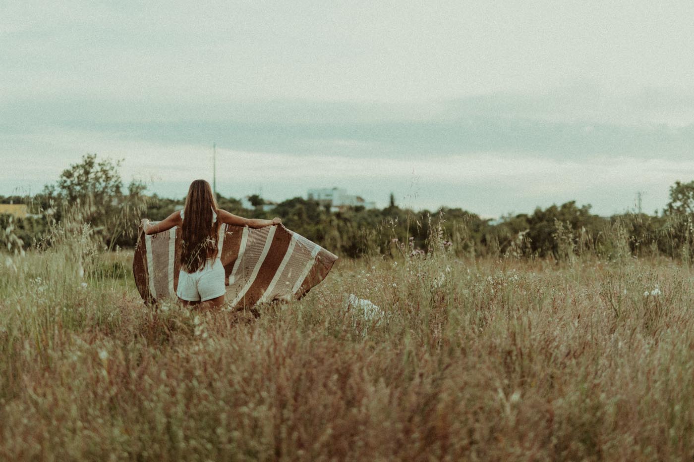 Une femme de dos, cheveux longs, marche au milieu d'un grand champ de hautes herbes en tenant une couverture à rayures.
