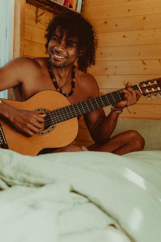 Un homme à la peau métisse et aux cheveux bouclés sourit en jouant de la guitare acoustique sur un lit.