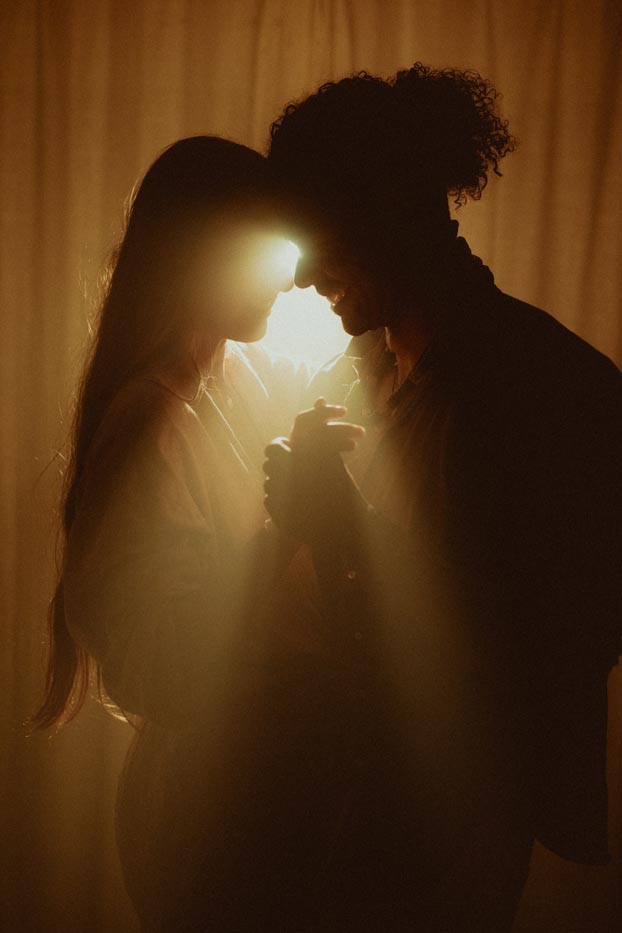 Silhouette en contre-jour d'un couple s'enlaçant tendrement face à face, baigné par une lumière chaude et brumeuse.