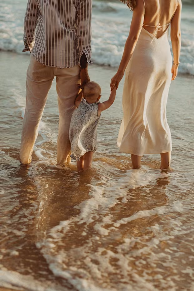 Deux adultes tiennent un bébé par la main dans l'eau de mer sur une plage au coucher du soleil.