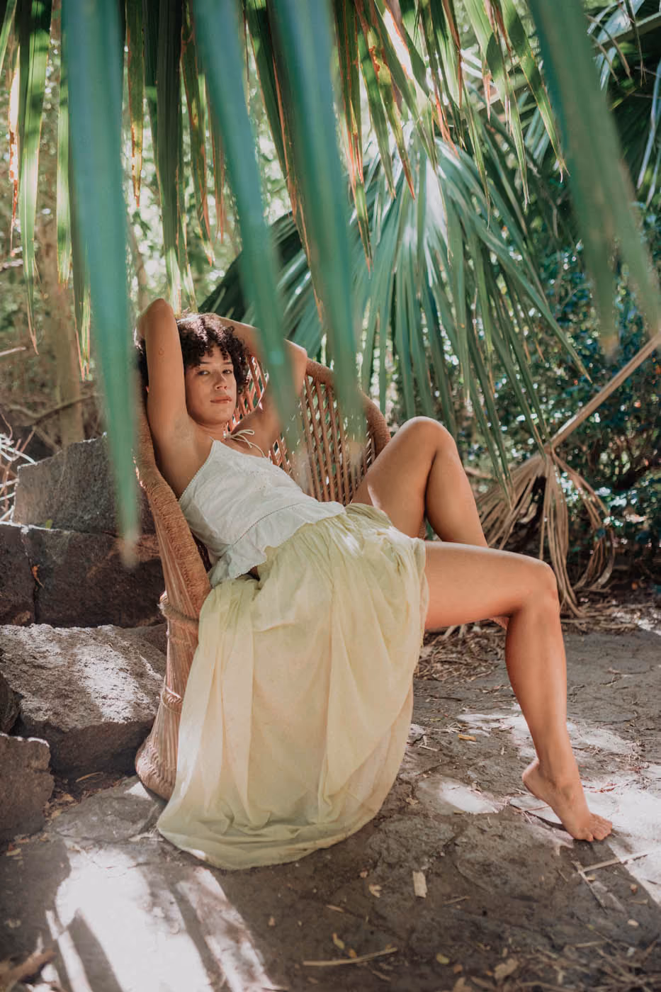 Une femme sur une chaise en rotin sous les feuilles de palmiers, vêtue d'un top blanc et d'une longue jupe jaune pastel.