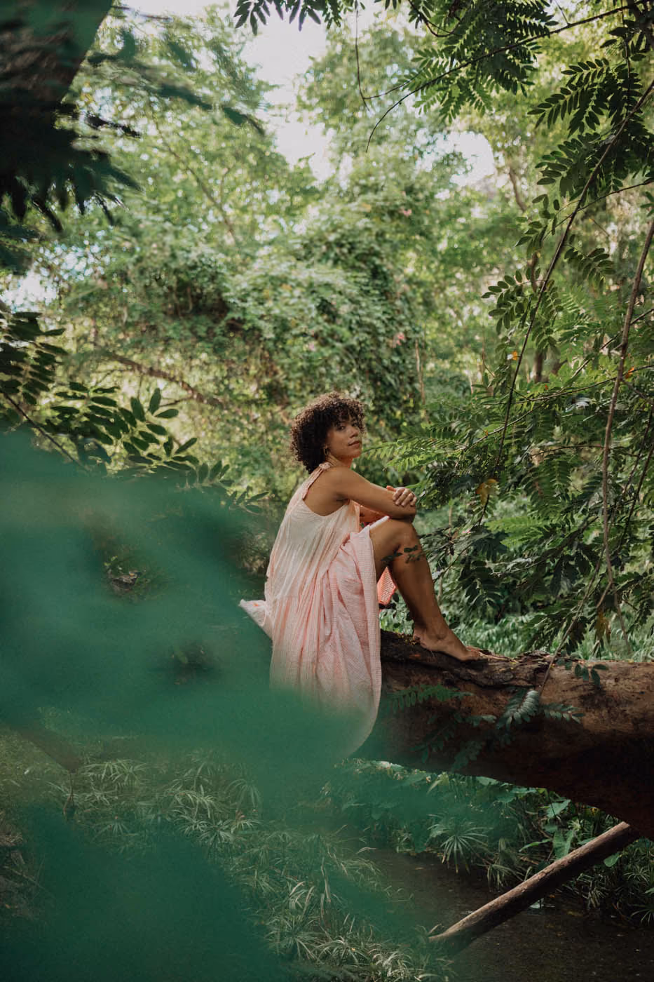 Portrait poétique d'une femme en robe rose assise sur un tronc d'arbre mort, cadrée à travers le flou artistique de feuilles vertes au premier plan.