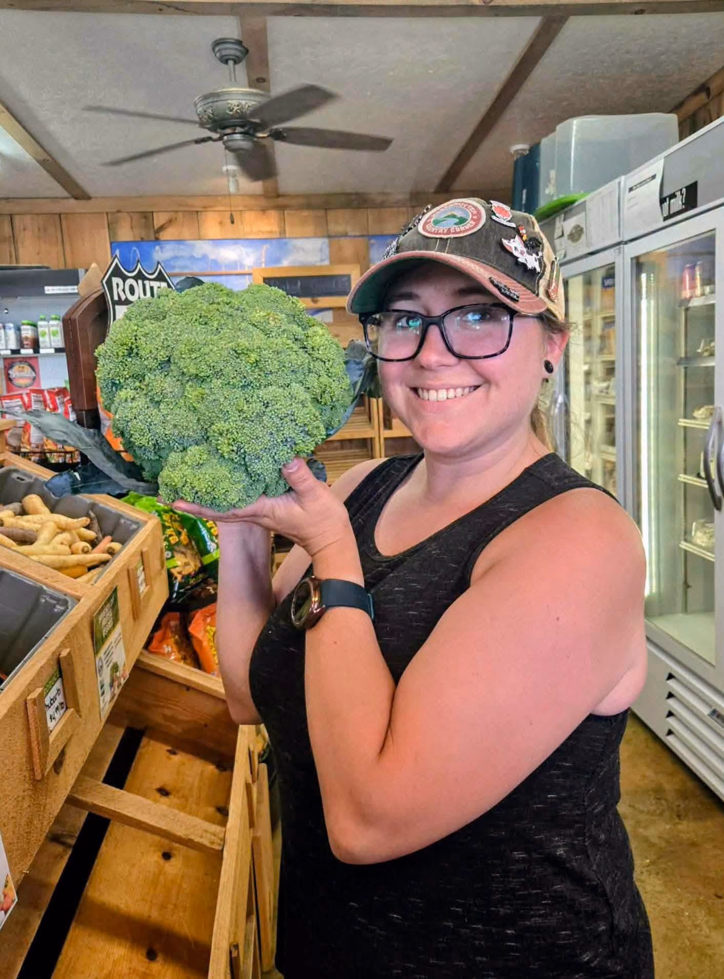 Smiling woman in cap and glasses holding large broccoli in a wooden produce market.