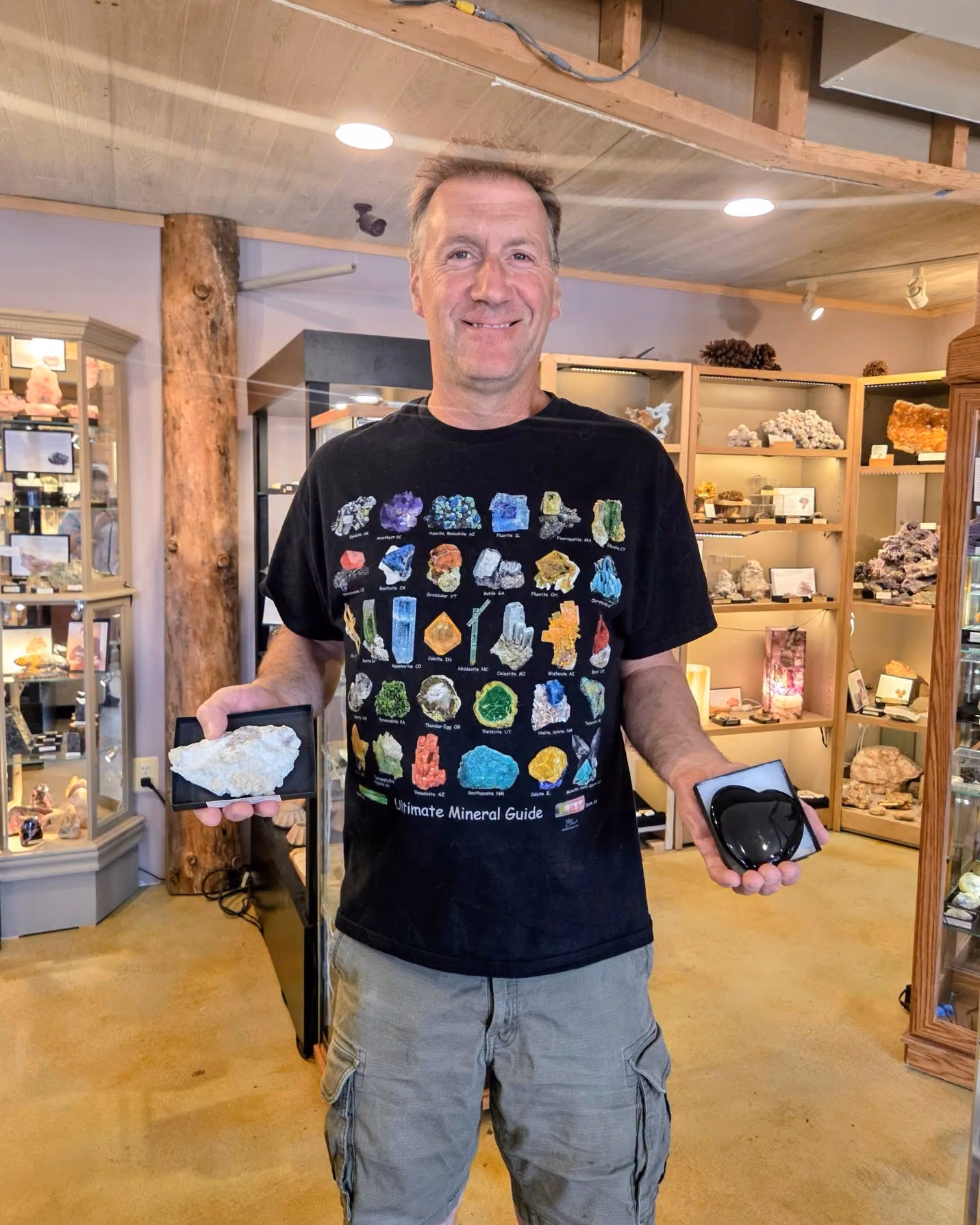 Man in mineral guide t-shirt standing in a mineral shop holding two different mineral specimens in each hand.