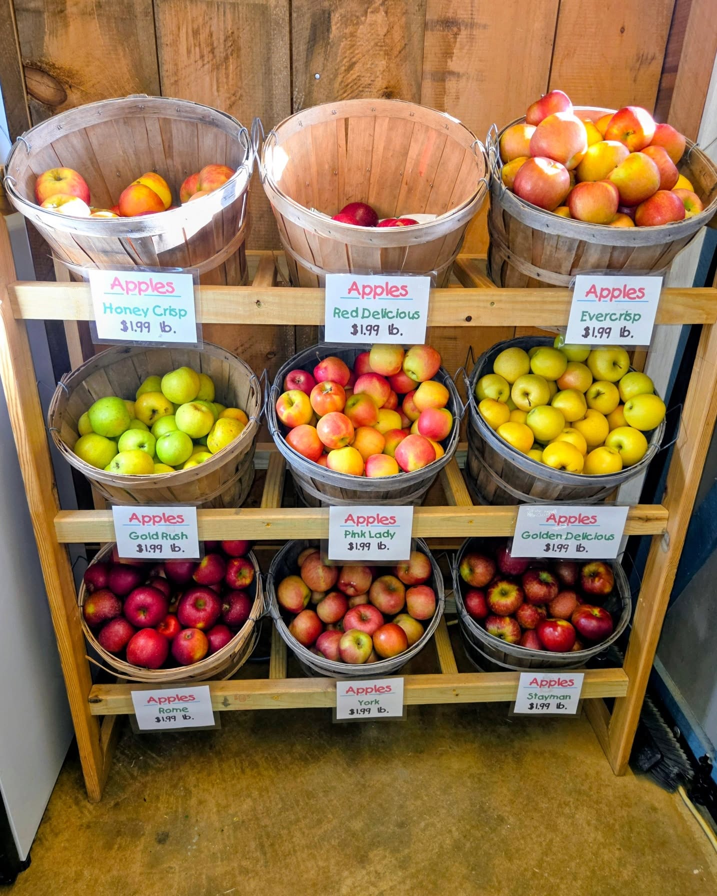 Wooden display racks holding nine baskets of various apple varieties with price tags.